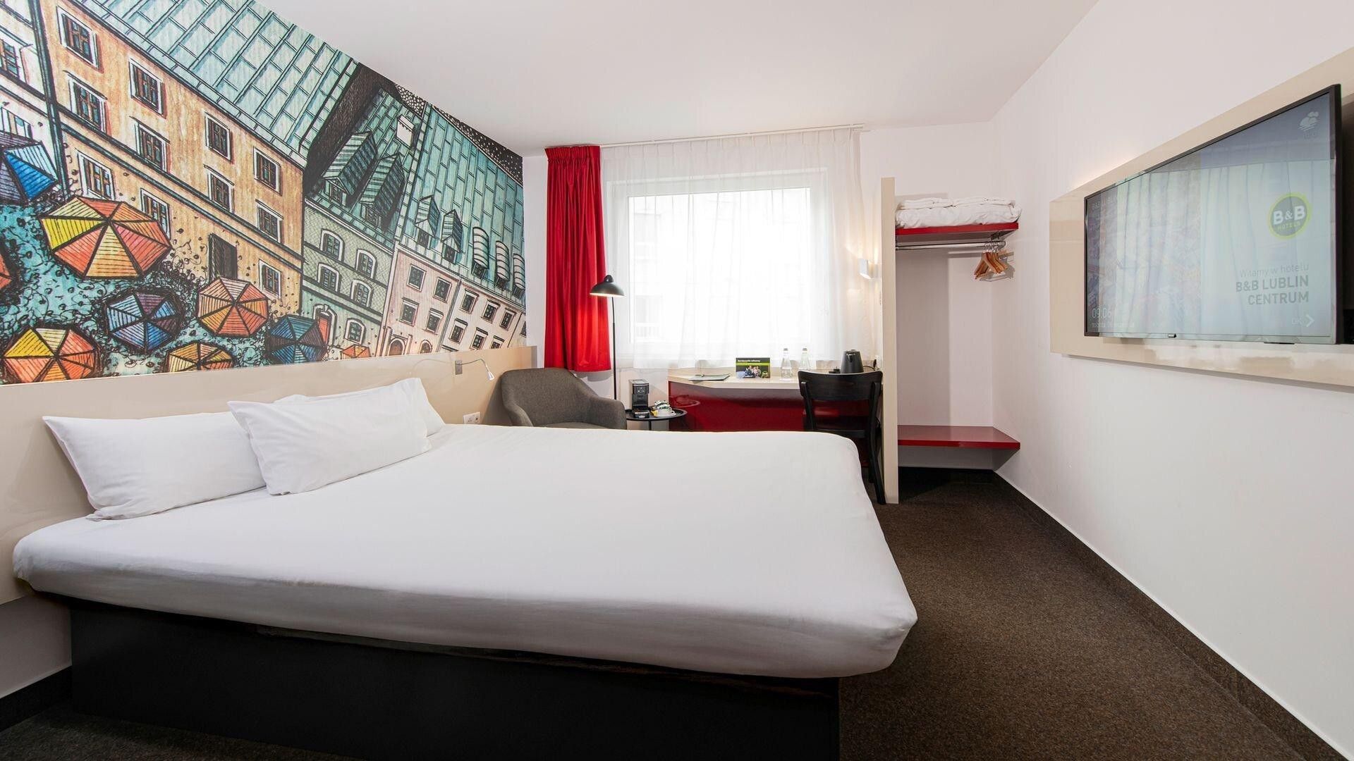A hotel room with a bed, a desk, a chair, a red curtain, and a wall with a colorful mural.