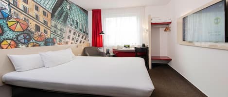 A hotel room with a bed, a desk, a chair, a red curtain, and a wall with a colorful mural.