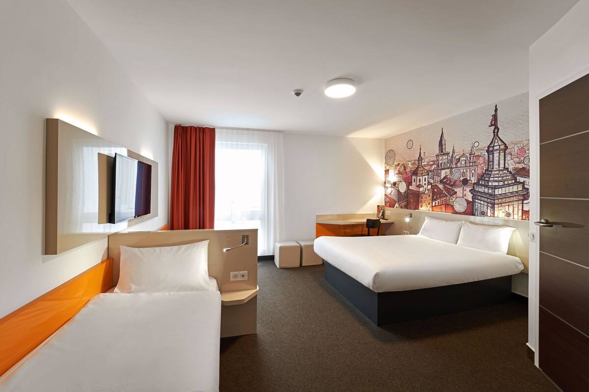 A hotel room with two beds, a desk, a chair, a TV, and a cityscape mural on the wall.
