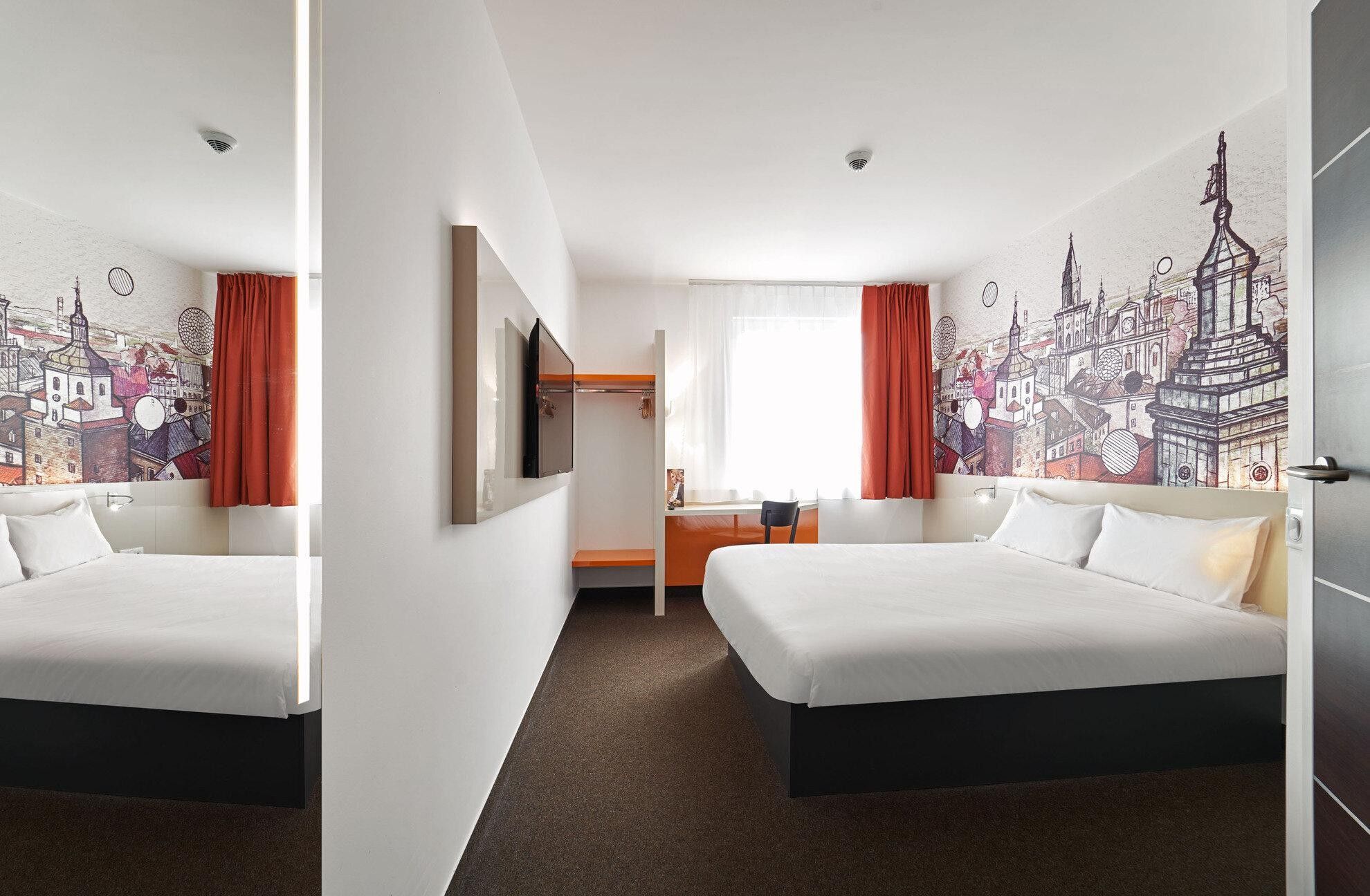 Habitación de hotel con dos camas, un escritorio y un mural de paisaje urbano en las paredes.