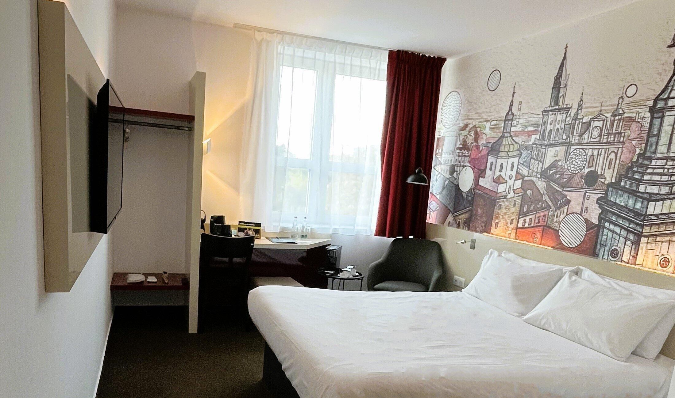 A hotel room with a bed, a desk, a chair, a TV, and a window with curtains.