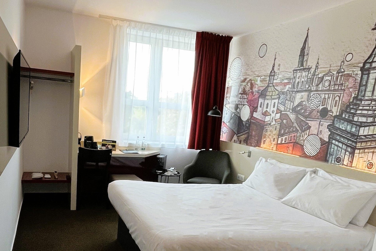 A hotel room with a bed, a desk, a chair, a TV, and a window with curtains.