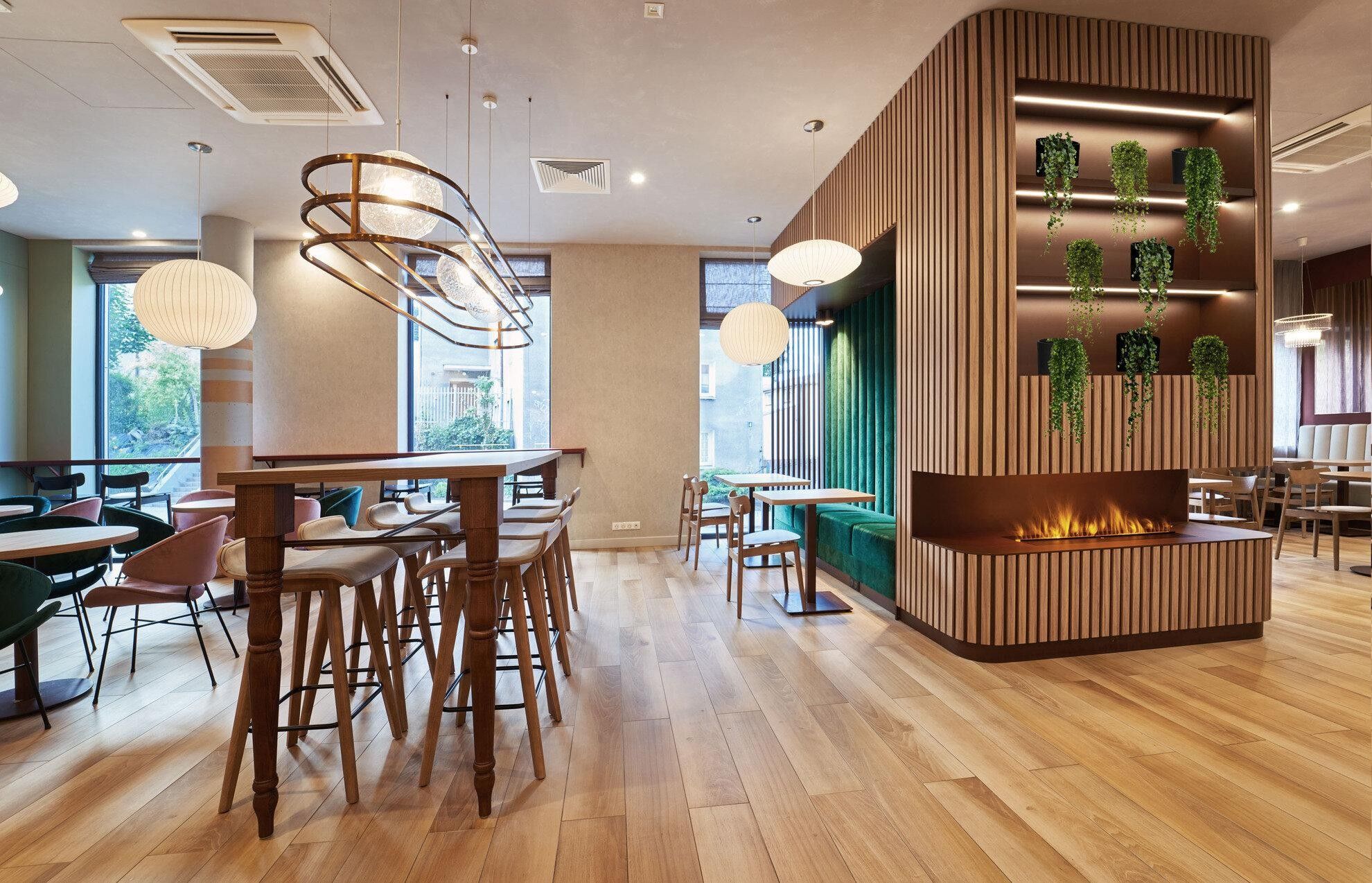 A modern interior with wooden flooring, round tables, and chairs, a central seating area with a built-in fireplace, and hanging plants.