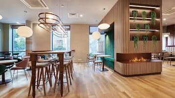 A modern interior with wooden flooring, round tables, and chairs, a central seating area with a built-in fireplace, and hanging plants.