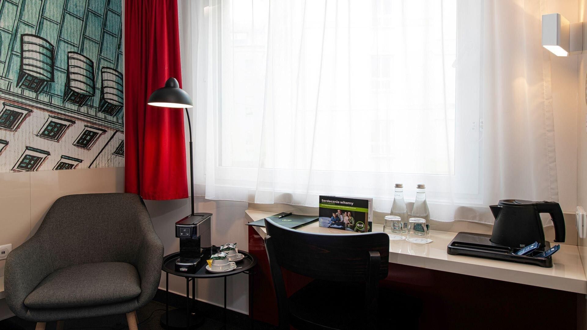A hotel room with a desk, chairs, a coffee maker, a lamp, and a window with curtains.