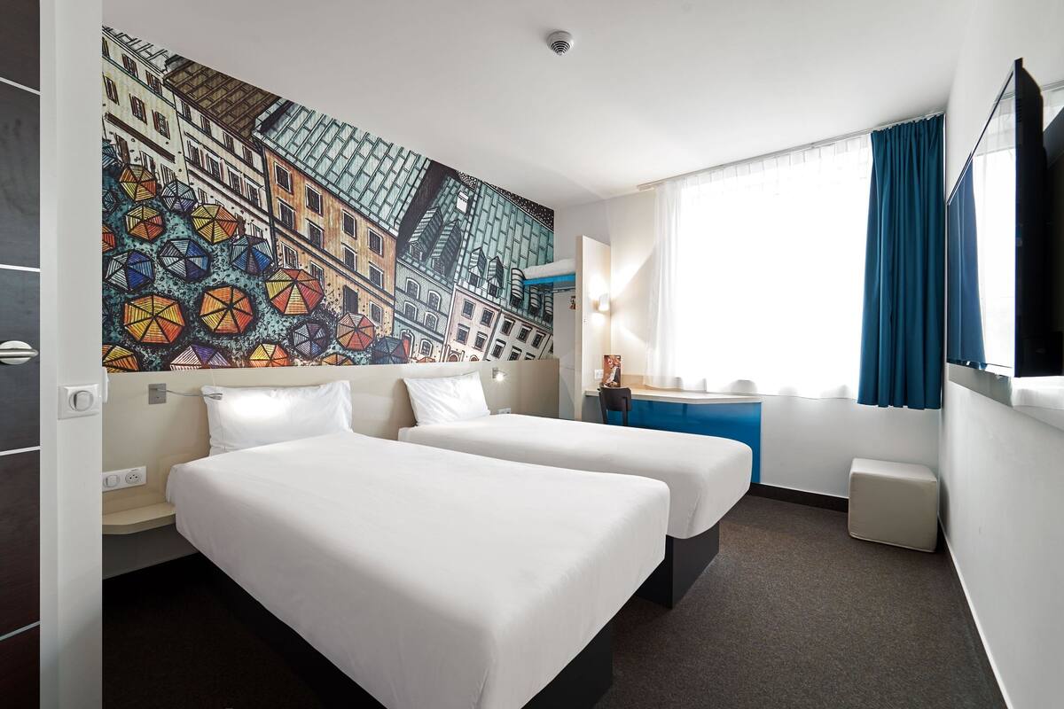 A hotel room with two beds, a desk, a chair, a window with curtains, and a wall mural.