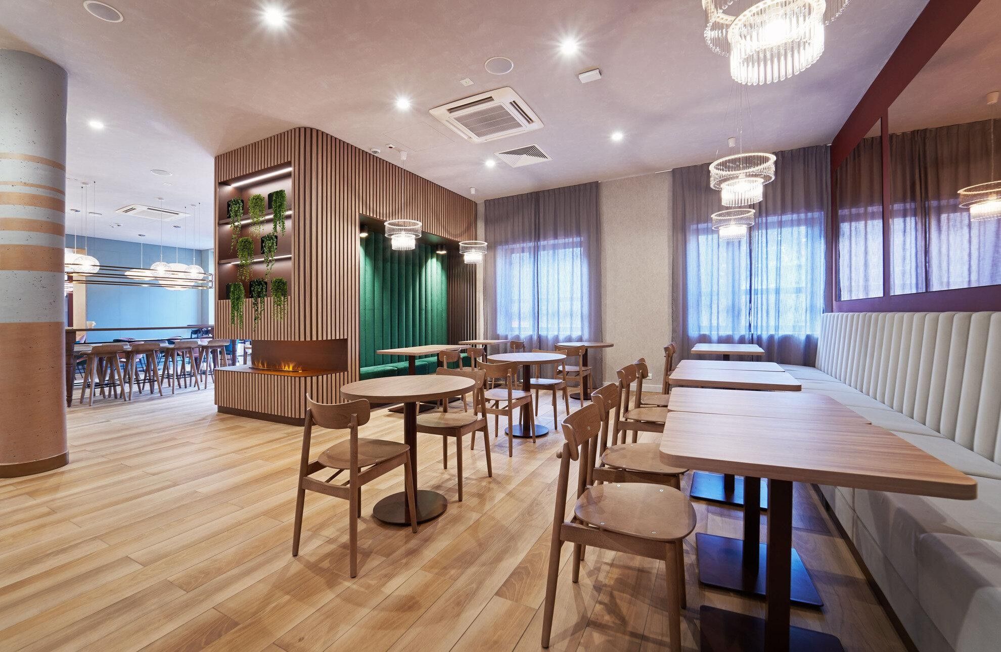 A modern restaurant interior with wooden flooring, round tables, and a green accent wall.