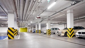 Un estacionamiento subterráneo de varios niveles con varios coches aparcados, conductos de ventilación y pilares con franjas amarillas y negras.