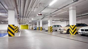 Garagem subterrânea com vários níveis, diversos carros estacionados, dutos de ventilação e defensas cilíndricas listradas em amarelo e preto.