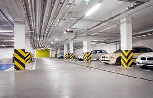 Garagem subterrânea com vários níveis, diversos carros estacionados, dutos de ventilação e defensas cilíndricas listradas em amarelo e preto.