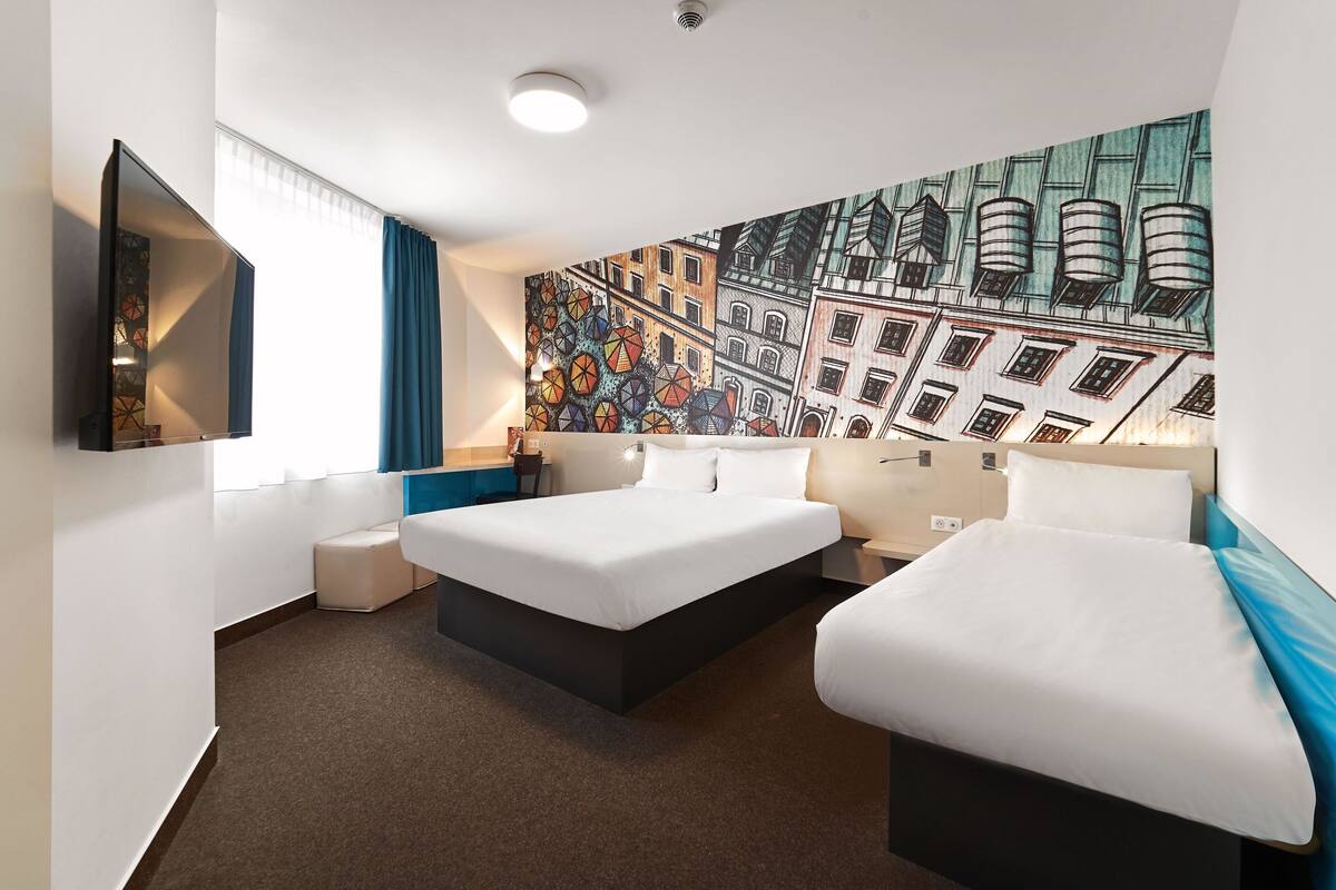 A hotel room with two beds, a large window with curtains, a wall-mounted TV, and a colorful mural.
