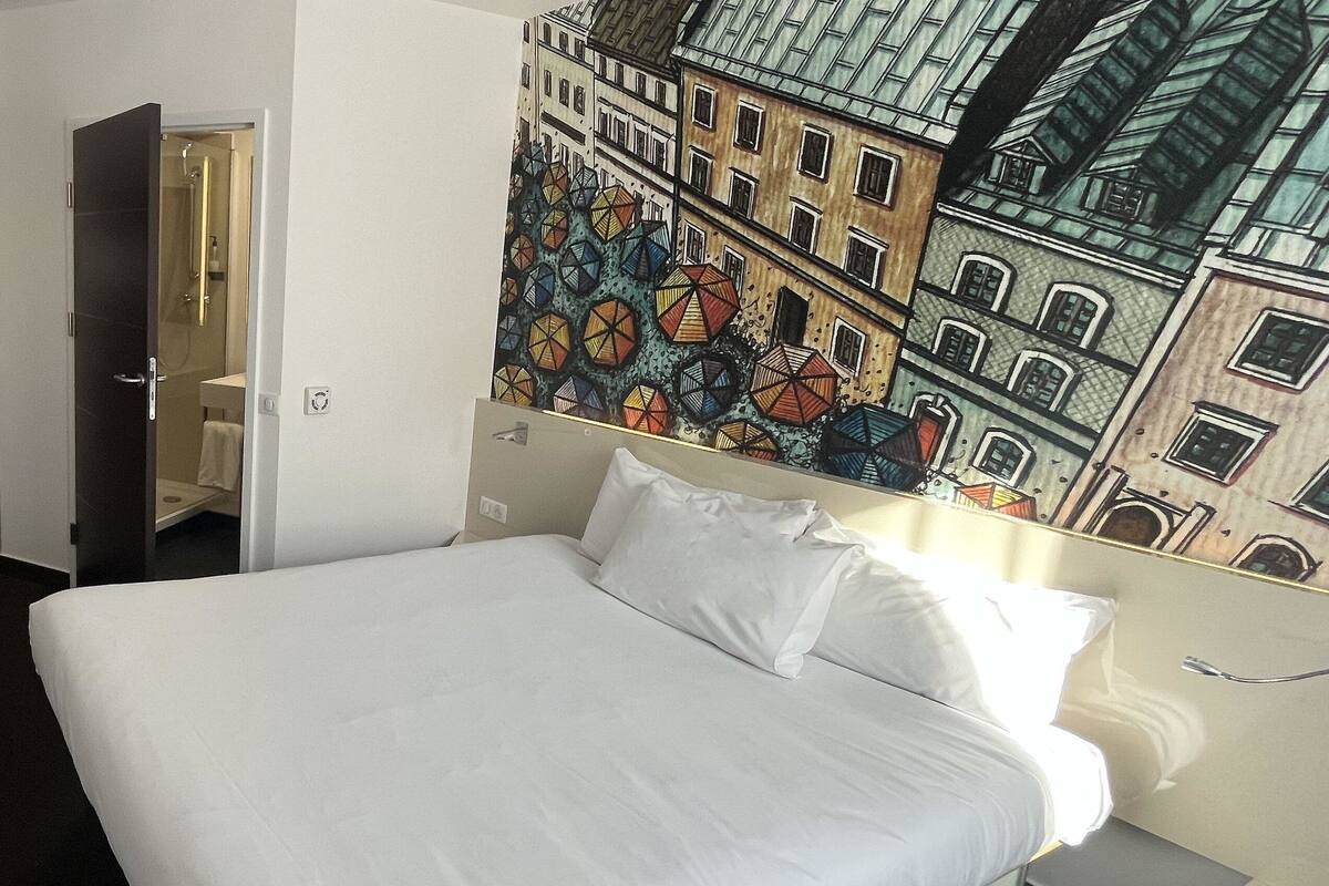 A hotel room with a large bed, a bathroom with a mirror, and a colorful mural on the wall.