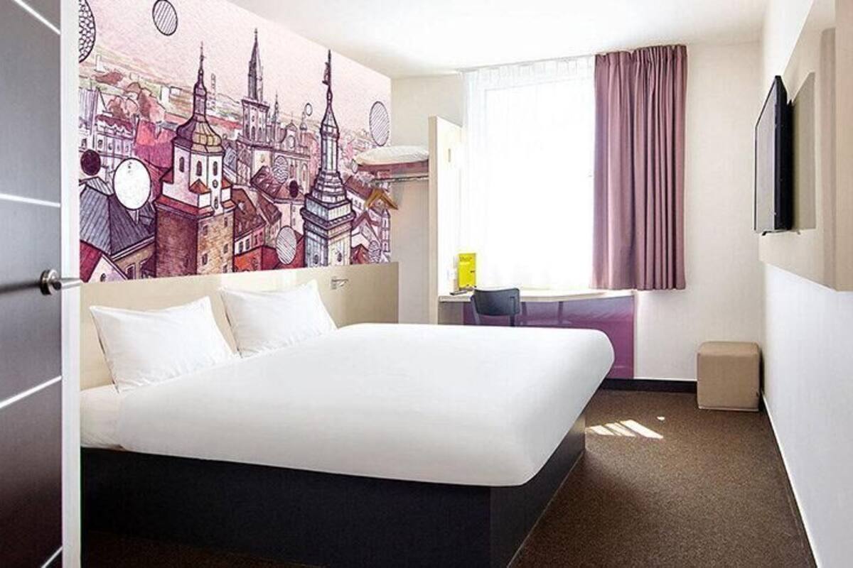 A hotel room with a large bed, a desk, a chair, a small table, a television, and a window with curtains.