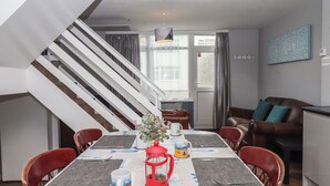 Un coin repas avec une table dressée pour quatre personnes, une lanterne rouge et un vase rempli de fleurs. On aperçoit un escalier au fond.