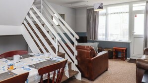 Un salon moderne avec un escalier, un coin repas avec une table dressée pour deux personnes et une grande fenêtre avec des rideaux.