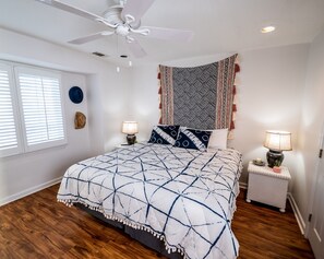 A bedroom with a bed, two bedside lamps, a ceiling fan, a window with blinds, a wall-mounted tapestry, and a small nightstand with a candle. - Atlantic Sunrise ~ Oceanfront, Beachfront, Pools (Tybee Island)