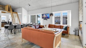 Een ruime woonkamer met een hoge plafond, een houten trap, een eethoek met grijze stoelen, een bruine leren bank en een aan de muur bevestigde televisie.