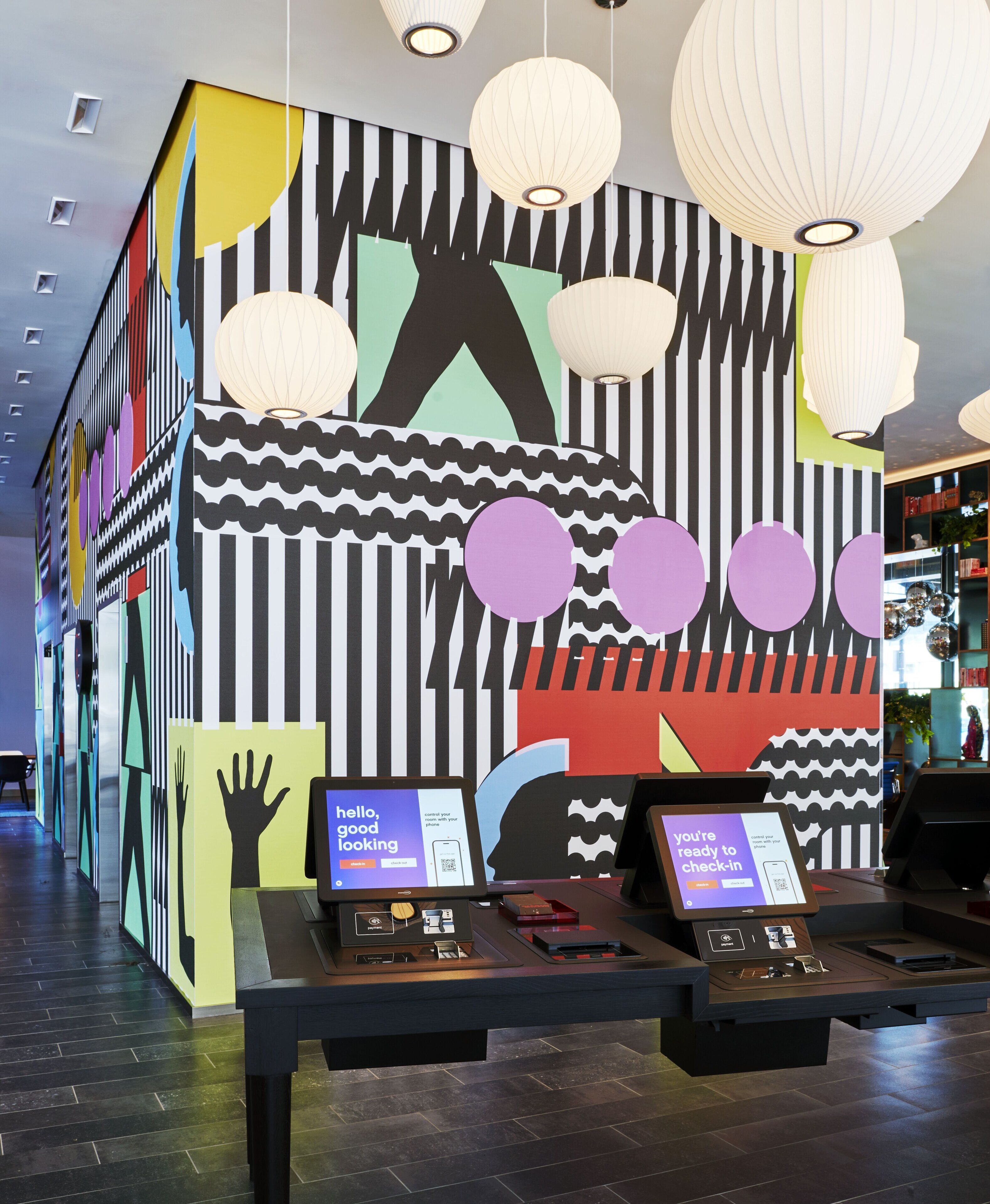 A modern interior with a check-in kiosk, a large abstract mural, and hanging paper lanterns.