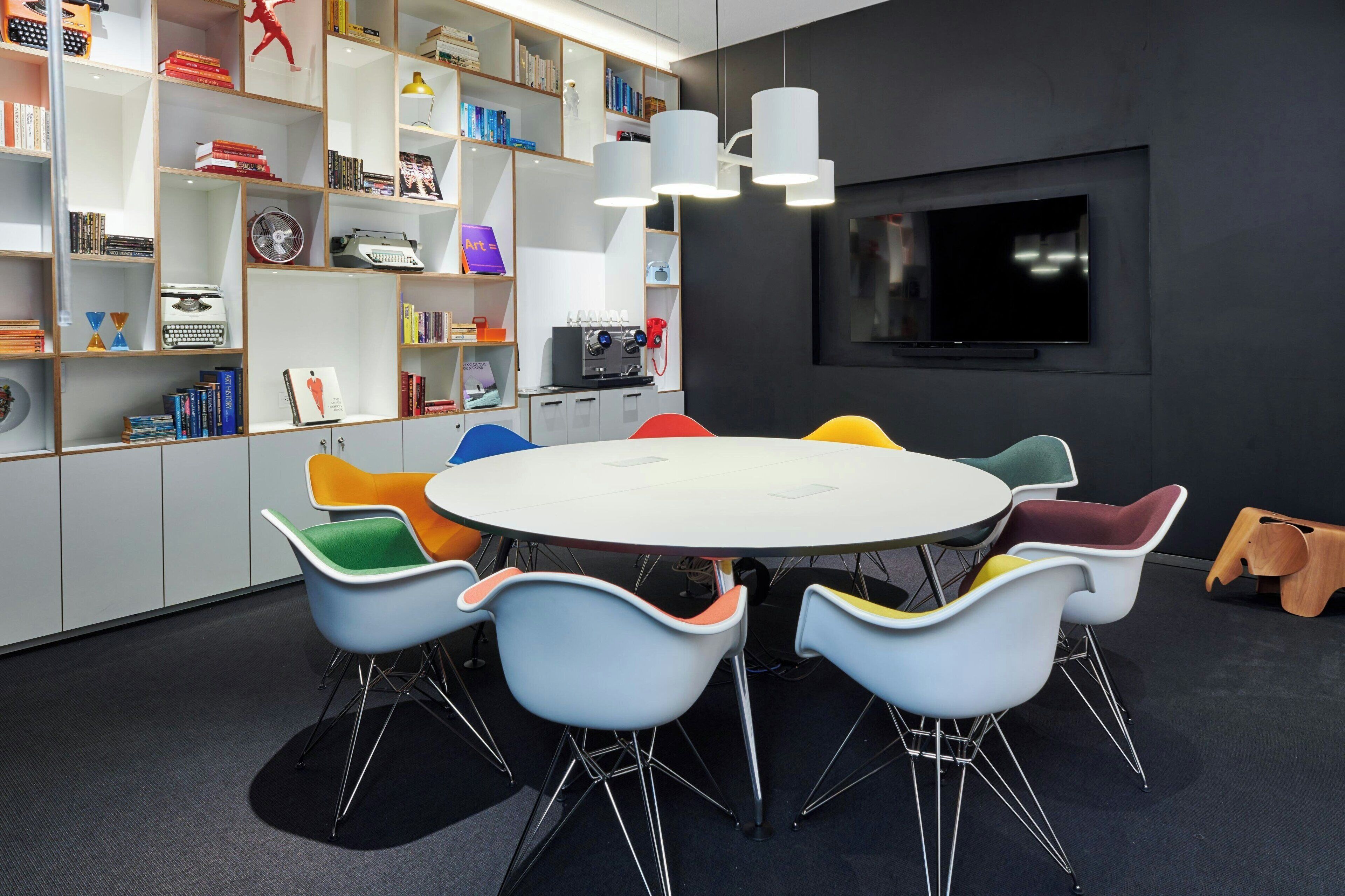 A modern meeting room with a round table, colorful chairs, a bookshelf, and a TV on the wall.