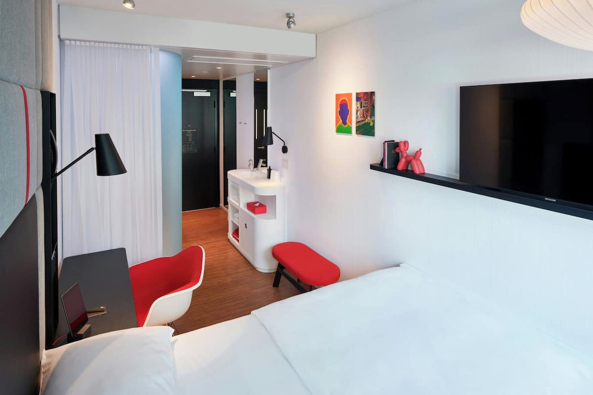 Ein modernes Hotelzimmer mit einem Bett, einem Schreibtisch mit einem Laptop, einem roten Sessel, einem kleinen Tisch, einem Fernseher und einem durchgegangenen Tür angedeuteten Badezimmer.