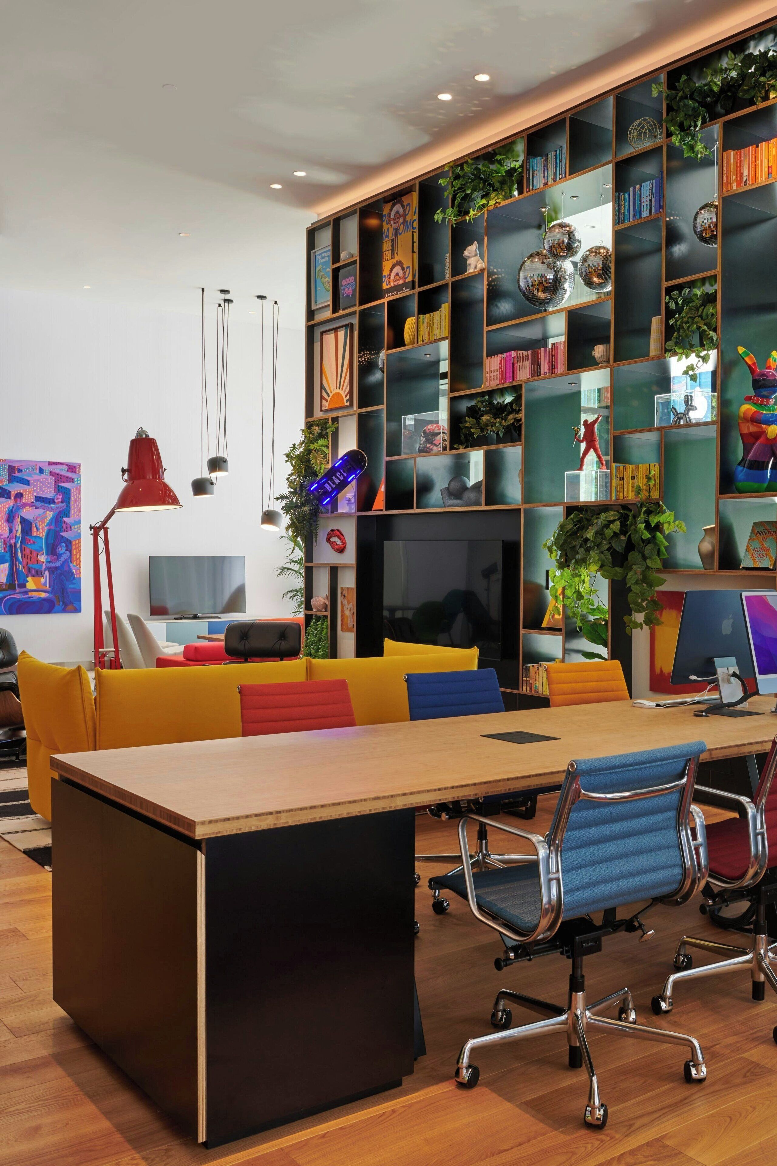 Un espacio de oficina moderno con una larga mesa de madera, sillas coloridas, una estantería llena de libros y objetos decorativos, y un televisor sobre un soporte.