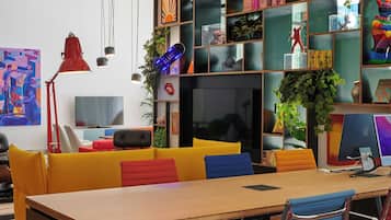 A modern office space with a long wooden table, colorful chairs, a bookshelf filled with books and decorative items, and a television on a stand.