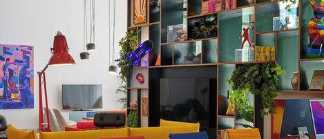 Un espacio de oficina moderno con una larga mesa de madera, sillas coloridas, una estantería llena de libros y objetos decorativos, y un televisor sobre un soporte.