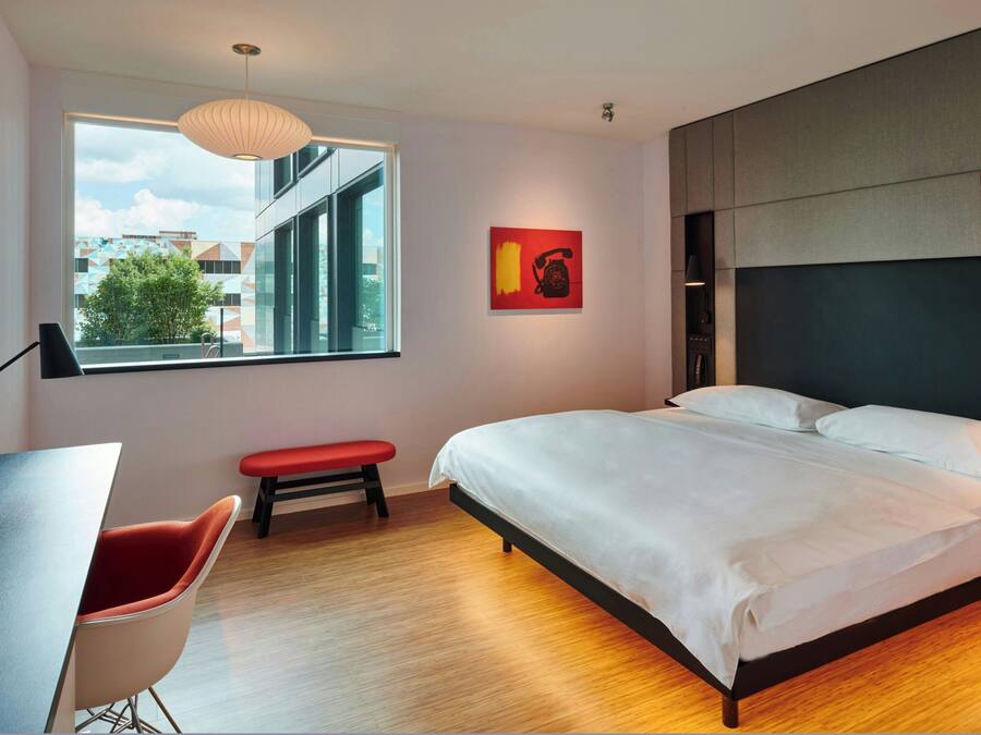 Een moderne slaapkamer met een groot bed, een bureau en een bankje. Er is een raam met uitzicht op gebouwen en bomen.