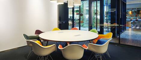 A conference room with a round table, colorful chairs, and large windows offering a view of the outdoors.