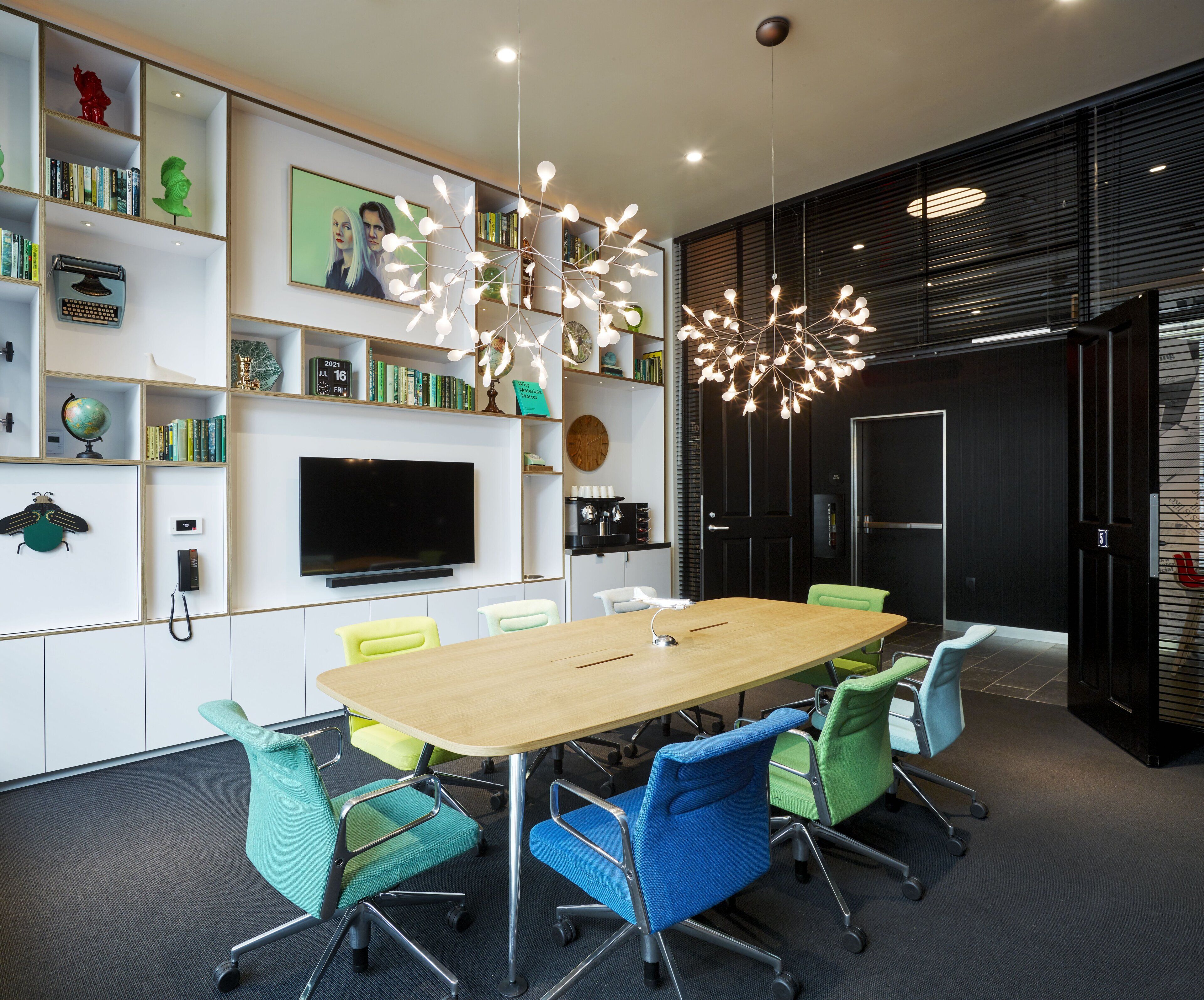 A modern conference room with a long table, colorful chairs, a large flat-screen TV, bookshelves, and a unique ceiling light fixture.