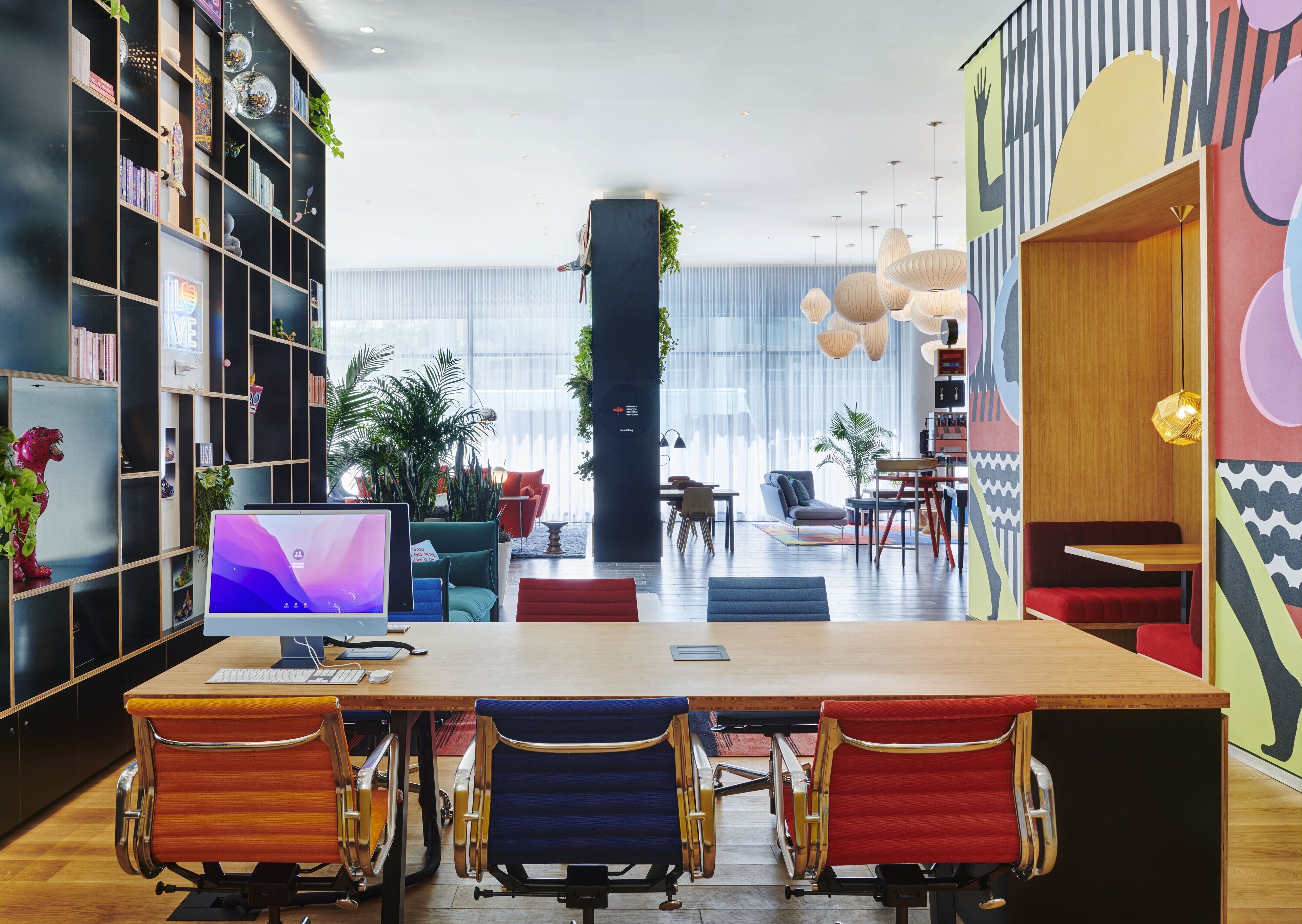 Un espace de bureau moderne avec une longue table, des chaises colorées et une grande bibliothèque.