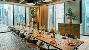 A long dining table set for a meal with chairs, large windows, and city views. - Hyatt Centric Melbourne (Melbourne)