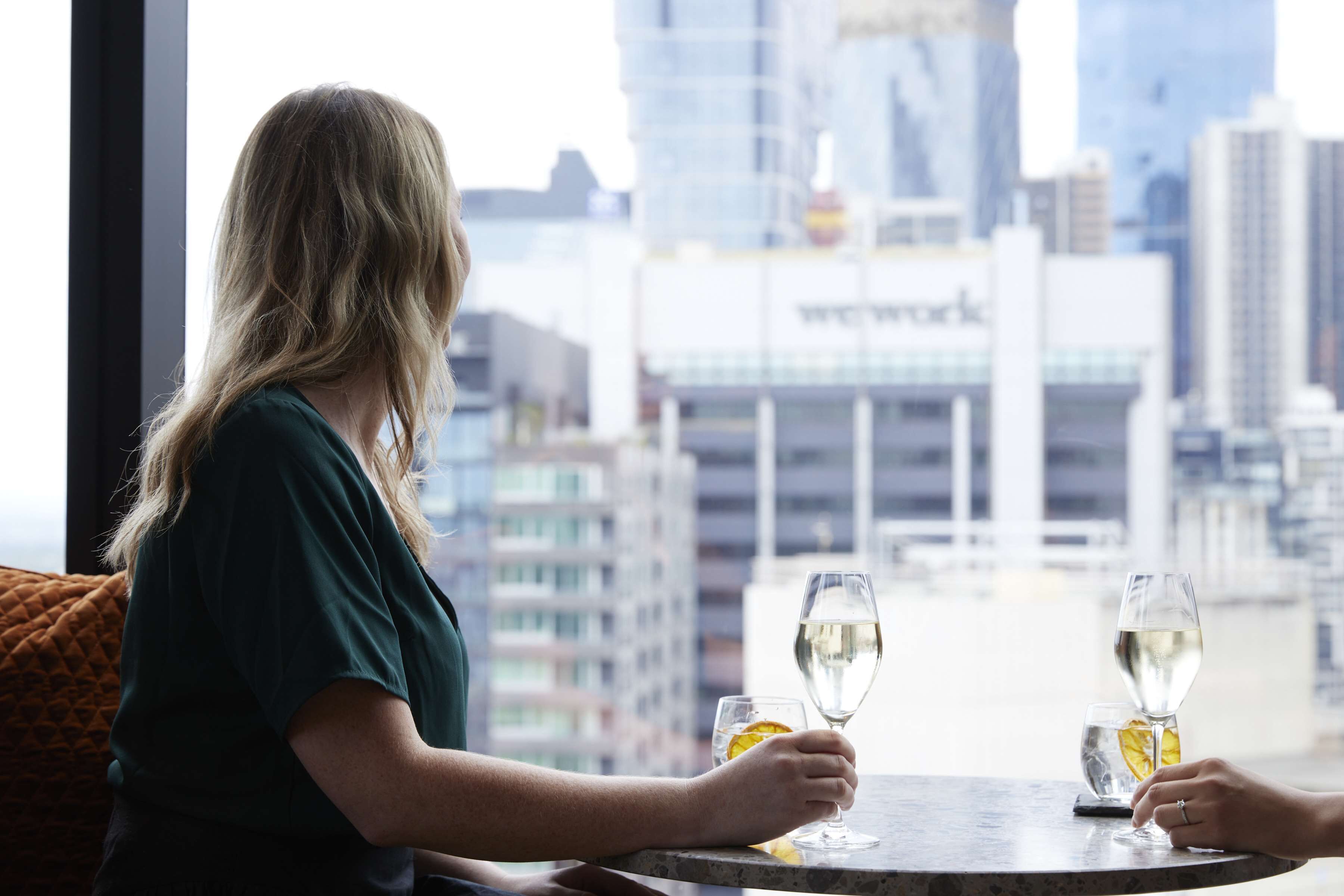 A person seated at a table with two glasses of white wine, one with an orange slice, in a modern urban setting with high-rise buildings in the background.