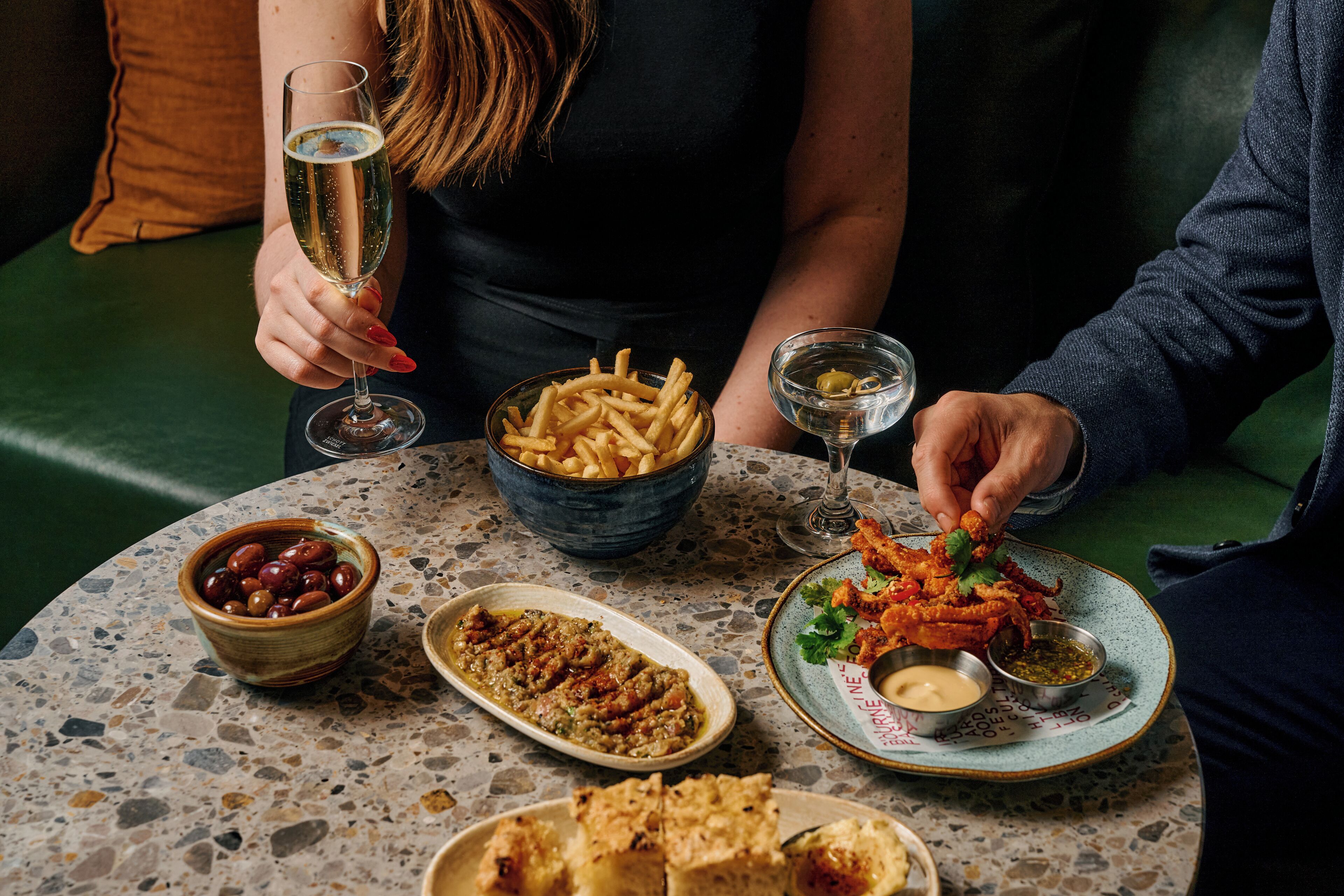 A table with various dishes, including a bowl of olives, a plate of fries, and a dish with a green sauce.