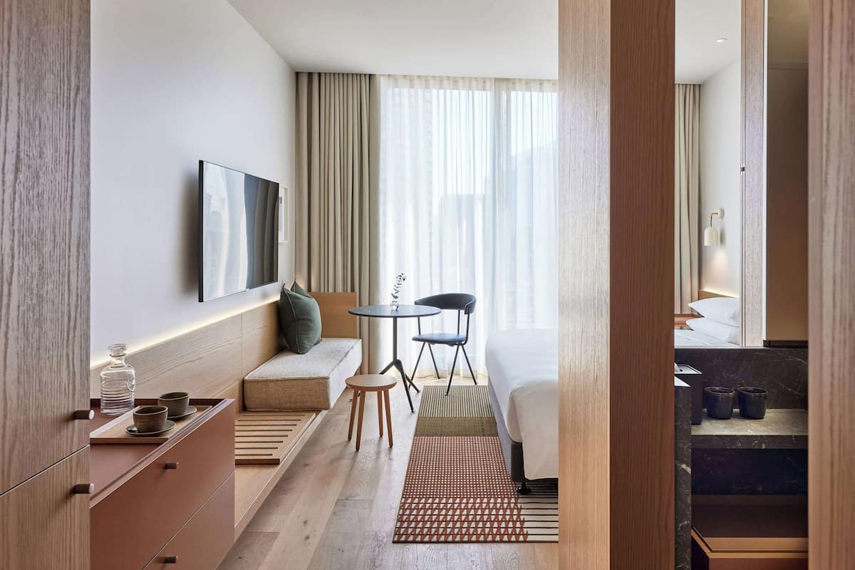 A modern hotel room with a sofa, a small table with chairs, a TV mounted on the wall, and a view of the city through large windows.