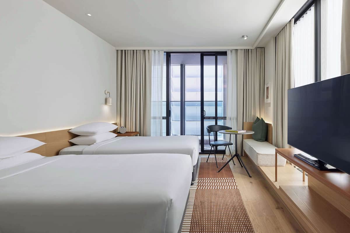 A hotel room with two beds, a TV, a small table, and a view of the city.