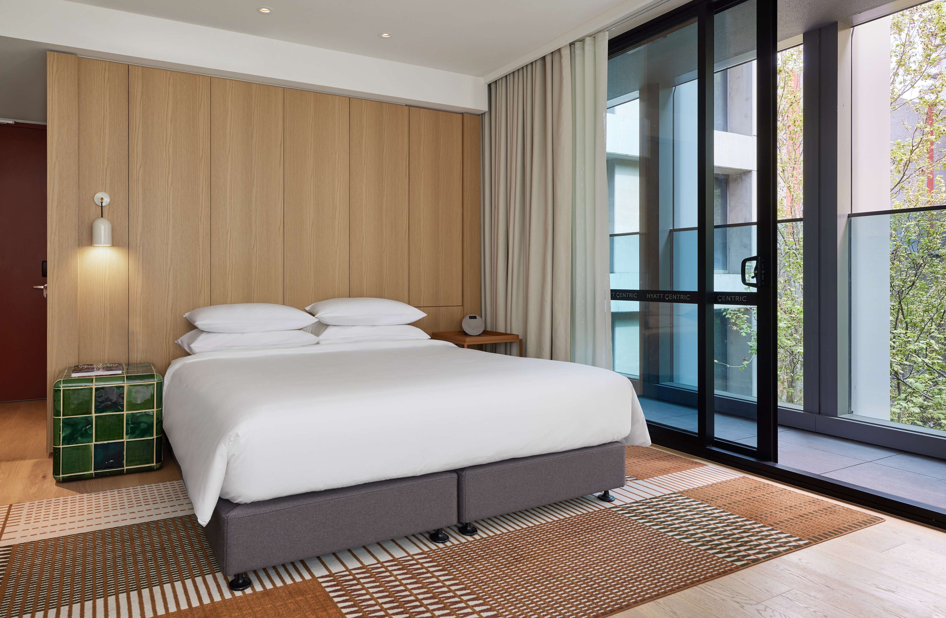 A modern hotel room with a large bed, a wooden panel wall, a large window with a view, and a small bedside table with a lamp.