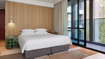 Una habitación de hotel moderna con una cama grande, un panel de pared de madera, un ventanal con vistas y una mesita de noche con lámpara.