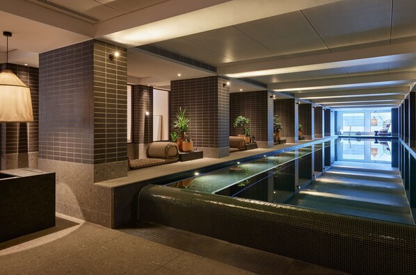 A modern indoor swimming pool with a long, narrow design, surrounded by seating areas and potted plants.