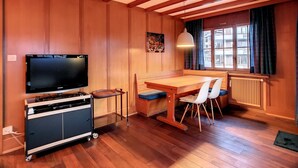 Quarto acolhedor com paredes e teto de madeira, uma mesa de jantar de madeira com cadeiras, uma televisão sobre um suporte e uma janela com cortinas.