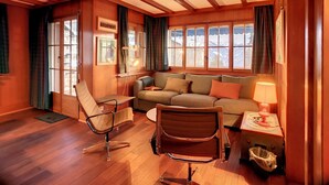 Quarto aconchegante com teto de madeira, um sofá, duas poltronas, uma mesinha com abajur e uma janela com cortinas.