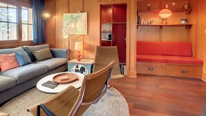 Uma sala de estar acolhedora com um sofá, uma poltrona, uma mesinha e um sofá vermelho. Há uma estante com livros e objetos decorativos, e um quadro na parede.