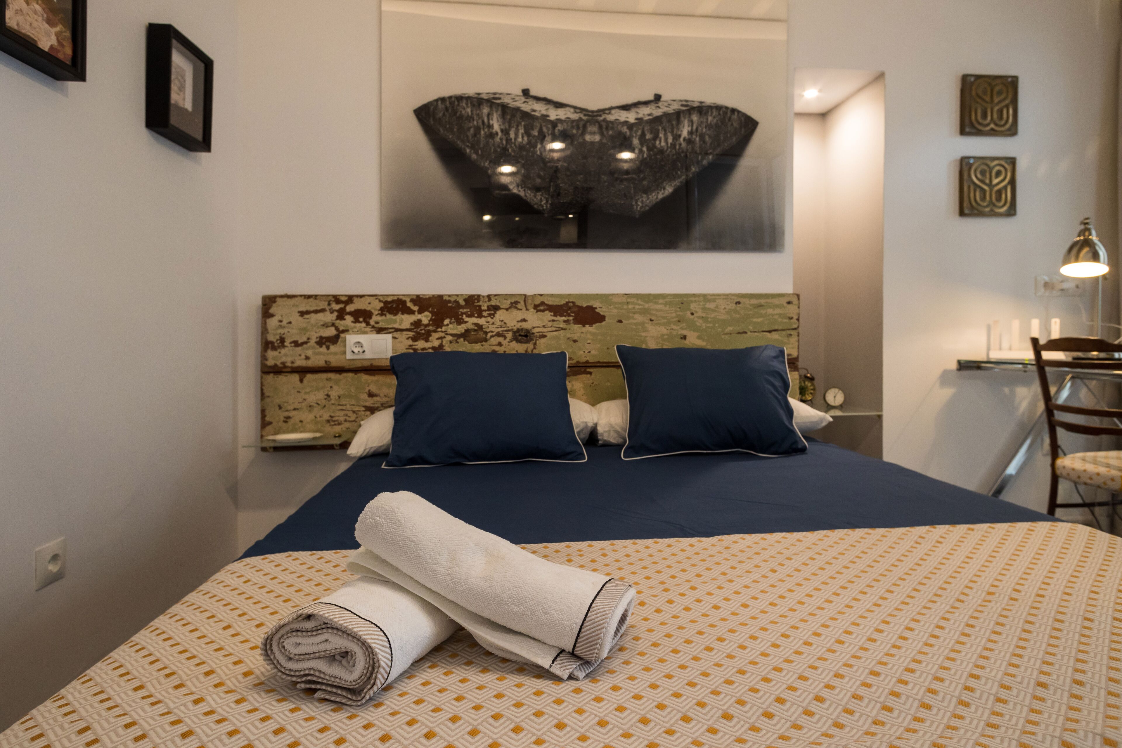 A hotel room with a bed, a headboard, a wall-mounted artwork, and a nightstand with a lamp.