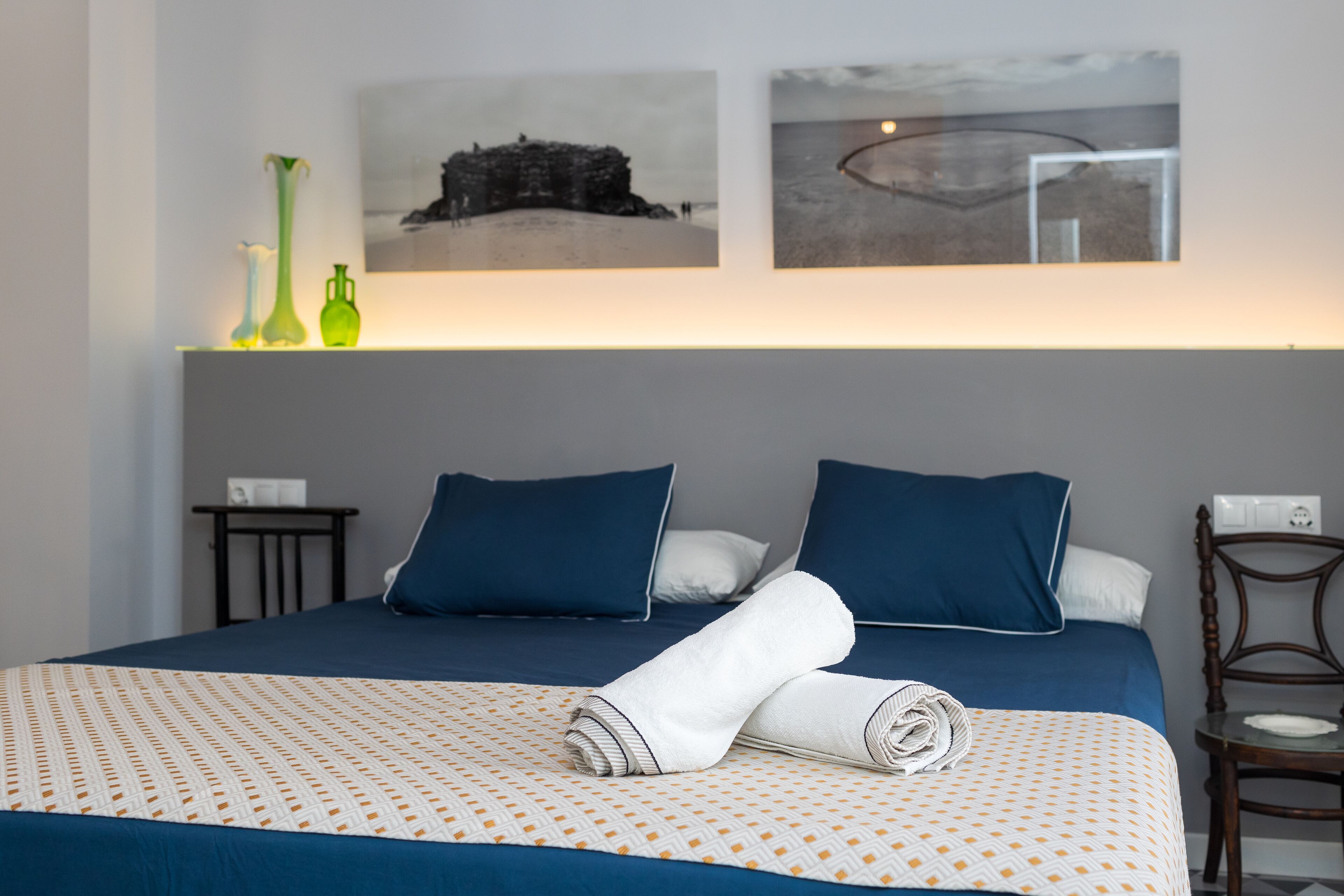 Un lit bien fait, avec une literie bleue et blanche, deux oreillers et une serviette pliée. Il y a deux petites tables, l’une avec un vase et l’autre avec une télécommande. La pièce présente un aménagement simple et épuré, avec un mur gris et un tableau encadré au-dessus du lit.