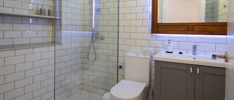 Un baño moderno con azulejos blancos tipo metro, ducha con cerramiento de vidrio, inodoro y tocador con espejo.