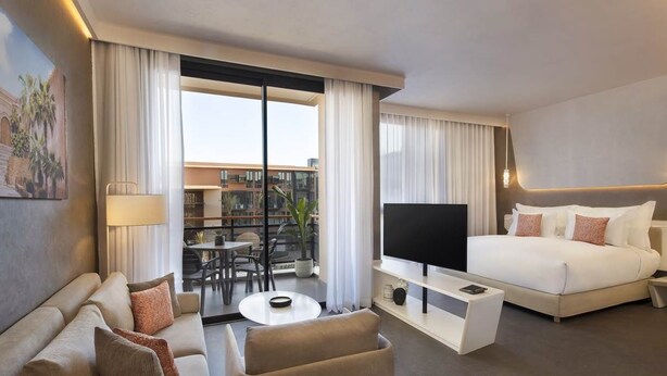 Una habitación de hotel moderna con una cama, televisión, sofá, mesa de centro y un balcón con vistas a edificios.