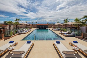 A rooftop pool area with lounge chairs and umbrellas, surrounded by palm trees and buildings.