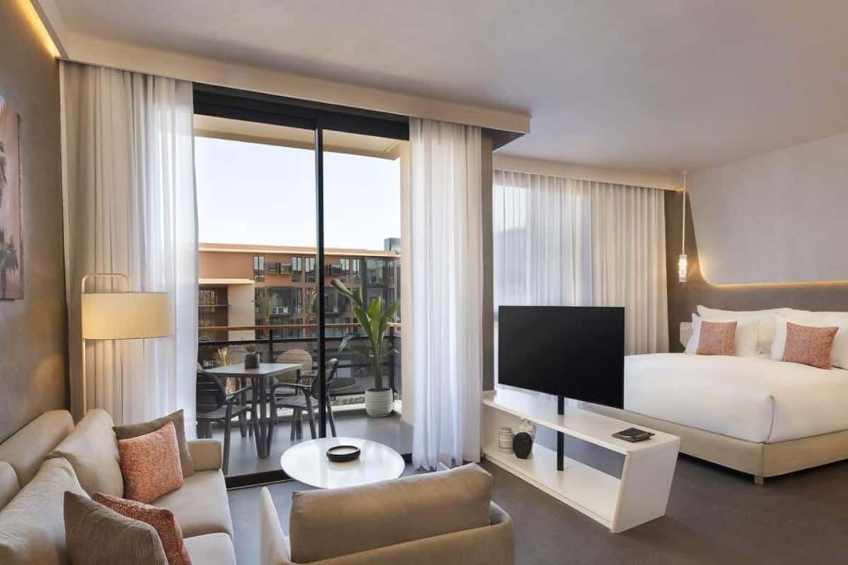 Ein modernes Hotelzimmer mit Bett, Fernseher, Sofa, Couchtisch und einem Balkon mit Blick auf Gebäude.