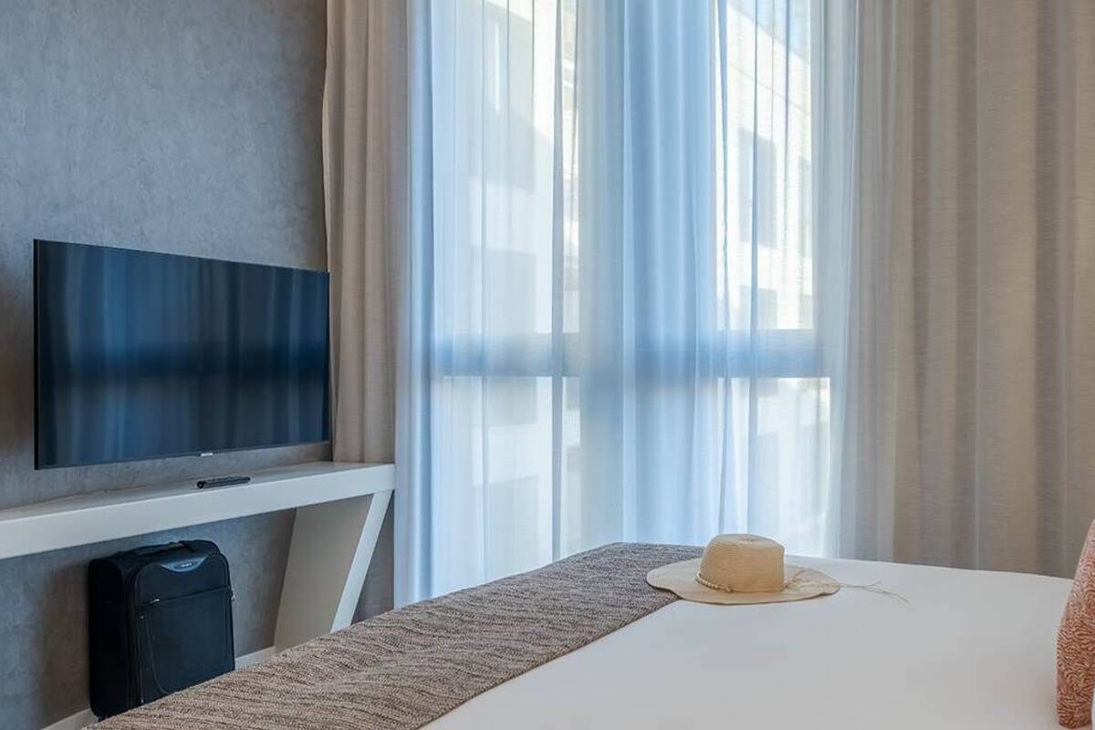 Hotelkamer met een flatscreen televisie, een bed met wit beddengoed, een hoed op het bed en dunne gordijnen.