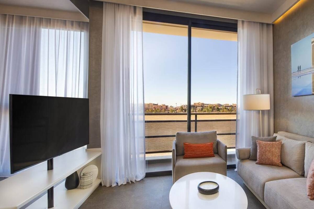 Ein Hotelzimmer mit großem Fernseher, einer Couch, einem Couchtisch und einem Balkon mit Blick auf die Stadt.